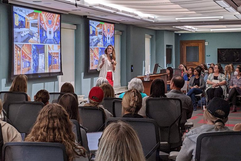 Photo of a woman with long hair wearing a white top and red skirt standing in front of a room of people presenting in front of projection screens.