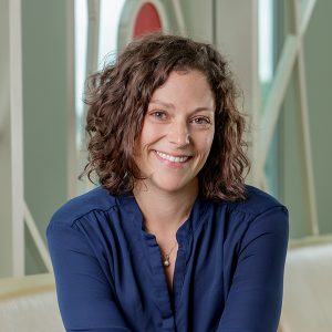Photo of a woman at bust length with shoulder length brown hair wearing a blue shirt seated and smiling at the viewer.