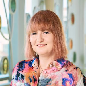 Photo of Anika Kozlowski, woman at bust length with a colorful shirt and light read hair smiling at the camera.