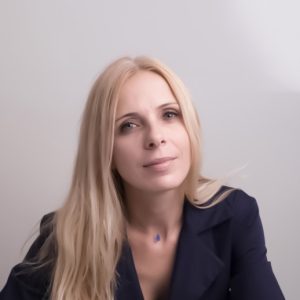 Photo of Monika Thadhani, woman and bust length with long, blonde hair and a dark blazer jacket smiling and looking at the camera.