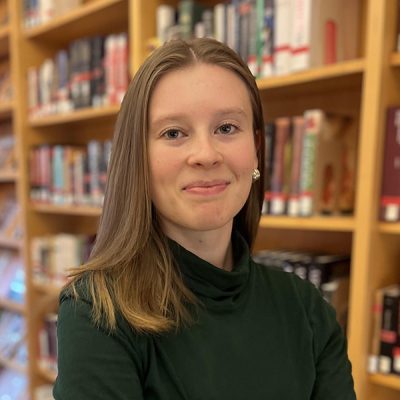 Photo of a woman at bust length standing in front of a bookcase. She has shoulder length light brown hair and is wearing a turtleneck