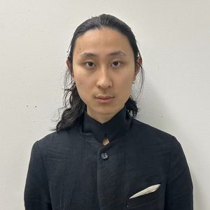 Photo of a man at bust length with long, black hair tied back, wearing a black top and looking at the camera.