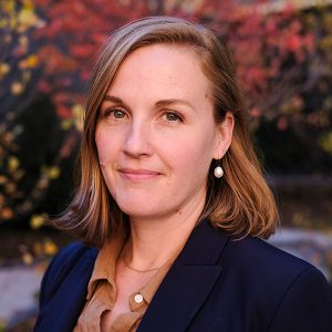 Photo of a woman at bust length wearing a blue blazer and brown shirt with shoulder length blonde hair smiling at the camera