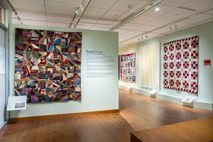 Installation of an art gallery with green walls showing several large quilts hanging on the walls and black text with a header that reads "Parallel Lines: Quilts and the American Landscape"