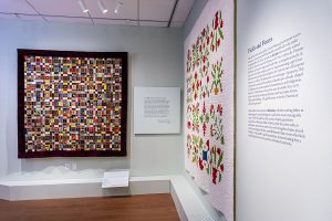 Photo of the corner of an art gallery with green walls with two large quilts hanging on the walls and labels in front of them below.