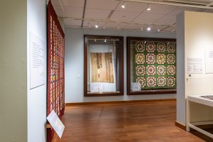 Photo of a gallery with several quilts hanging on the walls and a wood floor.