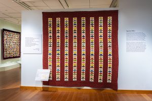 Photo of a large red quilt with colorful triangles in vertical rows hanging on a large blue wall.