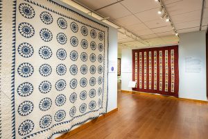 Photo of two large quilts on two separate walls, one that is white with blue circles and the other is red with colorful vertical rows on triangles.