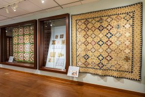 Photo of quilts in an art gallery. One is hanging on the wall to the right, and two are on view in cases to the left.