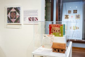 Photo of a case in an art gallery showing Lincoln logs and the historic box they come in.