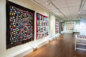 Photo of of several quilts hanging on a large, tan gallery wall with wood floors and mild lighting.