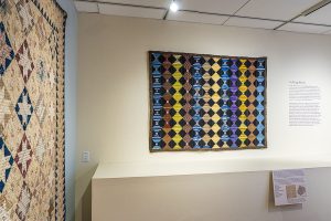 Photo of a quilt on a tan gallery wall. It is made of silt with a log cabin design in yellow, blue, and purple.