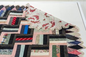 Detail of a quilt on a white table. The quilt has colorful silk strips and a lace edge.