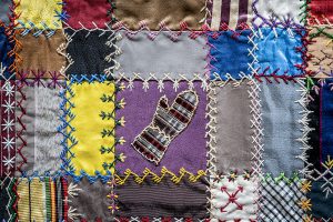 Detail of a patched quilt in in yellow, purple, grey, and brown with embroidery stitches and an embroidered mitten design in the center