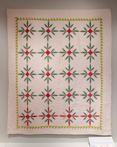 Detail of a white, red, and green floral quilt hanging on a wall in an art gallery.
