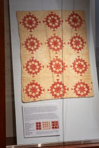 Photo of a red and white crib quilt on view in a glass case with a blue background.