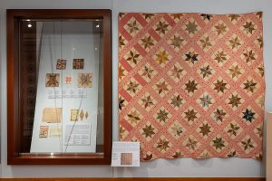 Photo of a quilt in an art gallery hanging on a wall with a case to the left showing fragments of quilts on a blue background.