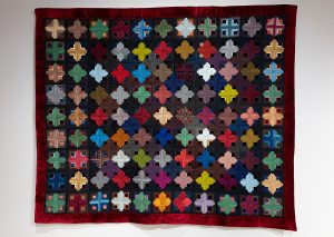 Photo of a quilt hanging on a tan wall. The quilt is made of silk with small, colorful crosses.