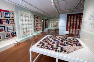 Photo of an art gallery showing several quilts hanging on the walls, and a silk quilt laying flat on a white table with an acrylic barrier.
