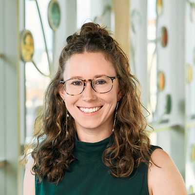 A white woman smiling, with brown, curly hair, and glasses, wearing a green sleeveless top and dangly beaded earrings.