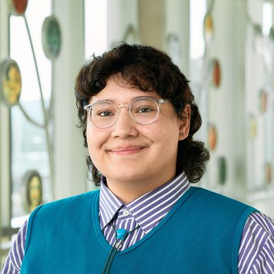 Hispanic person with short curly hair and glasses, wearing a purple striped button down, a cerulean vest, and a blue, heart-shaped bolo tie.
