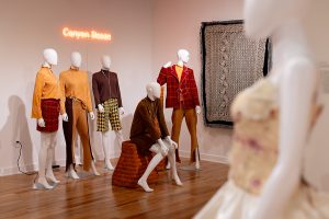 A photo of five mannequins display warm-toned, fall-colored garments. in front of a neon light that reads "Canyon Sleaze". A mannequin is featured in the right foreground, wearing a cream-colored dress.