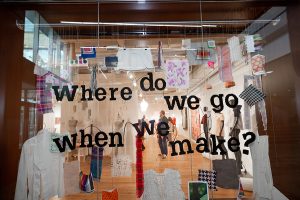 A close-up photo of the window display, which features fabric swatches and various garments suspended using wire. On the glass, the exhibition text reads "Where do we go when we make?"