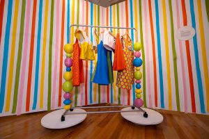 A clothing rack holds blue, yellow, orange, white, and green garments in front of a colorful striped background.