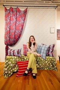 A person sits with their legs crossed on a couch with a green floral pattern, with a floral-patterned curtain behind them pulled to the left. On the couch sit four stripe-patterned pillows, with another pillow on the floor.