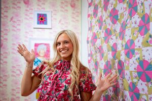 A person in pink clothing stands in front of two framed textiles, one featuring a starburst icon and the other a portrait of an older woman. To the right is a paper quilt made of blue, pink, white, and green pieces.