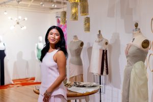 A person stands in a well-lit museum gallery, posing in front of three mannequins displaying beige garments and a round table with various fabrics laid atop.