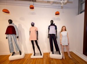 A person stands to the right of three mannequin displaying two tracksuit-like garments and one a short dress with a hat.