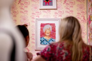 Two people stand in the foreground of the photo, viewing a framed textile portrait of an older woman that is displayed against a pink and yellow patterned wall.