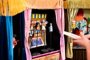 A close-up of a puppet box, with four figures on display stands. A pink curtain frames the box, with yellow fringe at the top of the box.