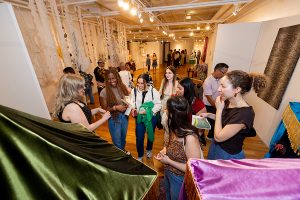 A group of seven people stand in front of three puppet boxes. Behind them are other people viewing various textiles throughout the gallery.