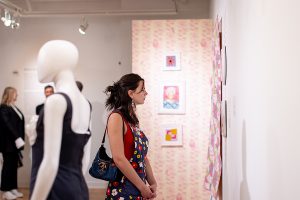 A person stands in a well-lit museum gallery, looking at textiles to the right. Behind them are three framed objects against a pink-patterned wall. In the foreground on the left is a mannequin displaying a sleeveless garment.
