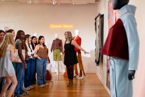 A person stands in front of an audience, looking at various textiles suspended to the right of the photo. Behind the speaker are two mannequins in fall-colors, with a neon sign that reads "Canyon Sleaze". In the foreground on the right of the photo there is a mannequin that displays a tracksuit.