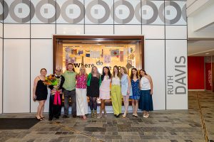 Photo of a group of people smiling and standing in front of an interior gallery window.