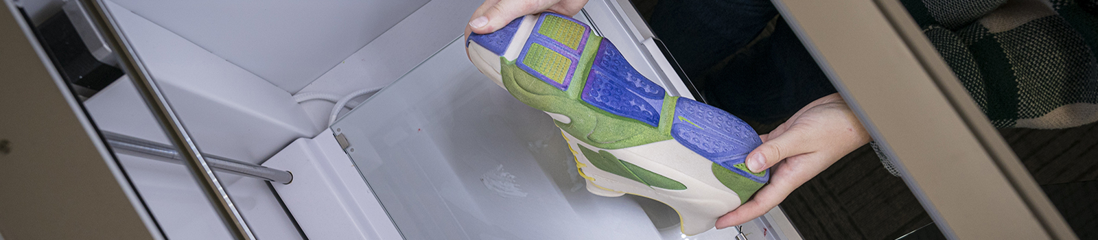 Photo of hands holding a 3D printed green and blue shoe inside of a 3D printer.