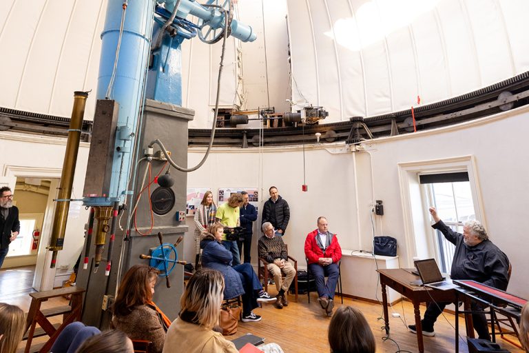 Photo of several people seated in an old observatory with a man in a black shirt seated at a table in front gesturing towards the dome of the building.