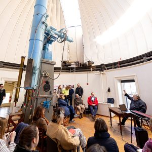 Photo of several people seated in an old observatory with a man in a black shirt seated at a table in front gesturing towards the dome of the building.