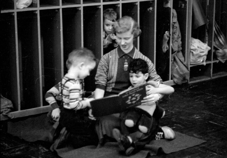 "Student teacher, Marilyn Nelson, sitting on the floor and reading to Mark Weinberg. Also listening are Frank Effland and Eddie Grant. Mark is holding a stuffed animal, and behind them, a child is hiding in a coat cubbyhole."