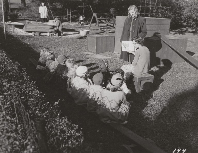 "Teachers reading to children at the Nursery School outside in the playground."