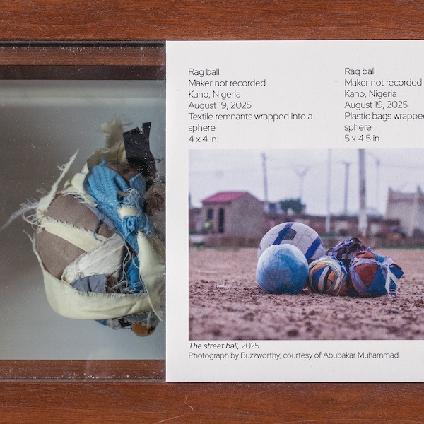 Close up photo with a ball made out of scraps of fabric within a wooden and glass display case. To the left of the ball in the case is a museum panel with information on the "rag ball" and its description.